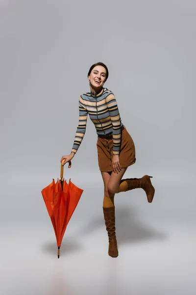 Full length of smiling young woman in retro autumnal outfit and boots posing with umbrella on grey — Stock Photo