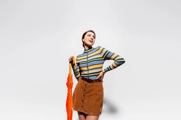 Femme heureuse en col roulé rayé et jupe posant à la main sur la hanche et tenant parapluie orange sur gris — Photo de stock