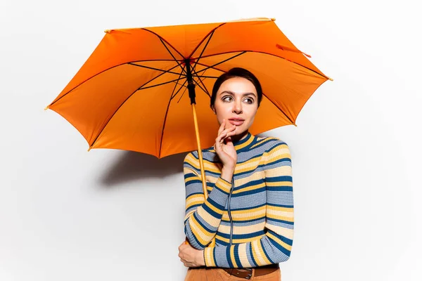 Mulher sonhadora em gola alta listrada em pé sob guarda-chuva laranja no branco — Fotografia de Stock