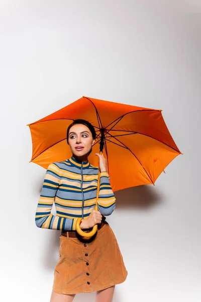 Брюнетка в полосатой водолазке и юбке, стоящая под оранжевым зонтиком на сером — стоковое фото