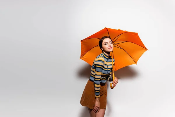 Giovane donna in dolcevita a righe e gonna in posa con ombrello arancione su grigio — Foto stock