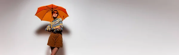 Morena mujer joven en cuello alto rayado y falda de pie bajo paraguas naranja en gris, bandera - foto de stock