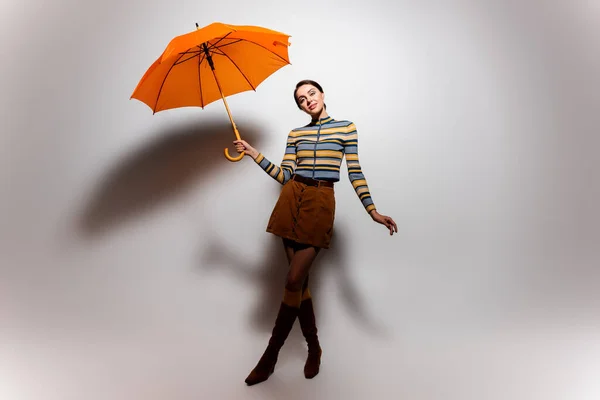 Full length of cheerful young woman in striped turtleneck and skirt posing with umbrella on grey — Stock Photo