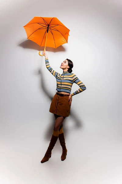 Piena lunghezza di donna allegra a righe collo alto e gonna in posa con ombrello su grigio — Foto stock