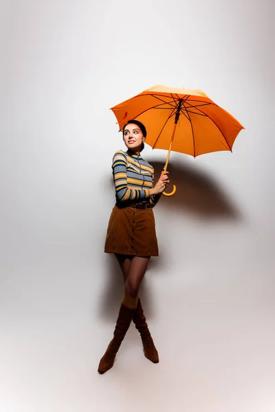 Volle Länge der lächelnden jungen Frau in gestreiftem Rollkragen und Rock posiert mit orangefarbenem Regenschirm auf grau — Stockfoto