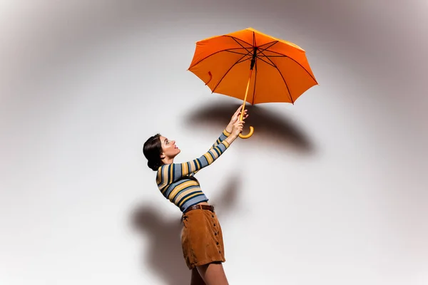 Vista lateral da morena jovem em gola alta listrada e saia posando com guarda-chuva laranja no cinza — Fotografia de Stock