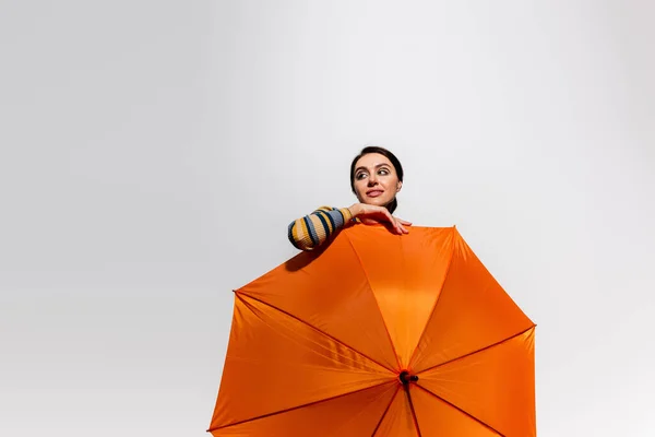 Verträumte junge Frau posiert mit leuchtend orangefarbenem Regenschirm isoliert auf grau — Stockfoto