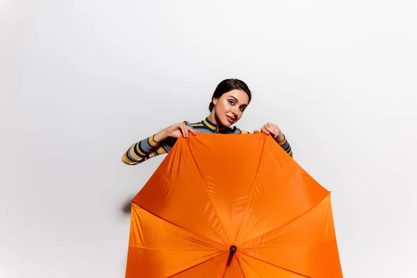 Morena joven en cuello alto rayado sosteniendo paraguas naranja sobre gris - foto de stock