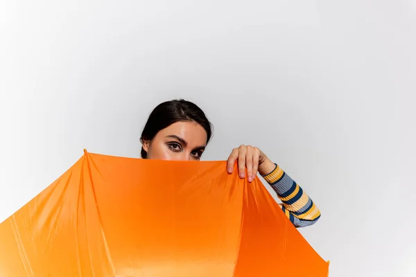 Brünette junge Frau verdeckt Gesicht mit orangefarbenem Regenschirm isoliert auf grau — Stockfoto