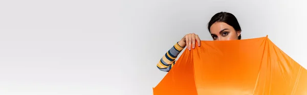 Brunette young woman covering face with orange umbrella isolated on grey, banner — Stock Photo