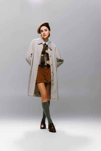 Full length of young woman in beret and vintage trench coat posing while looking at camera on grey — Stock Photo