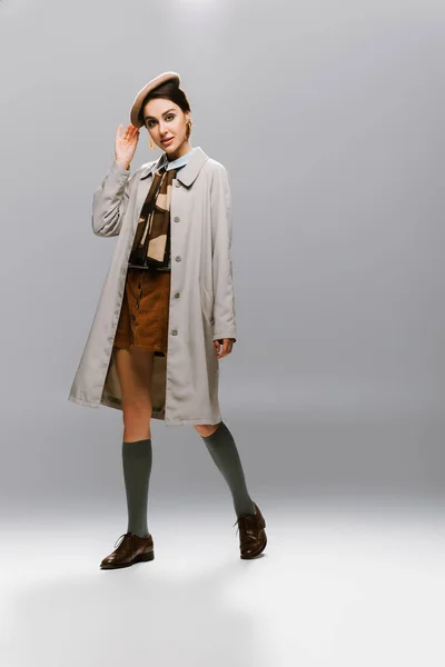 Повна довжина елегантної молодої жінки в автентичному тренч пальто регулювання берета на сірому — стокове фото