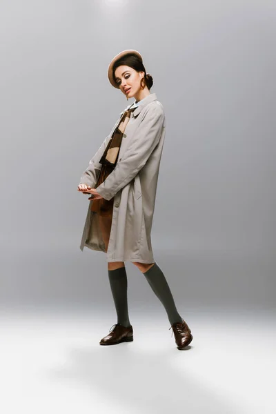 Volle Länge der eleganten jungen Frau in Baskenmütze und stilvollem Trenchcoat posiert auf grau — Stockfoto