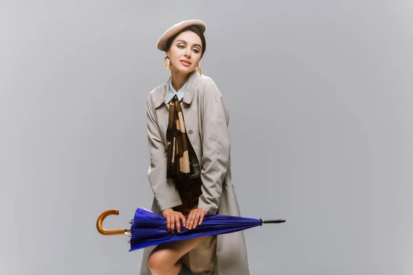 Элегантная молодая женщина в берете и плаще позирует с зонтиком изолированы на сером — стоковое фото
