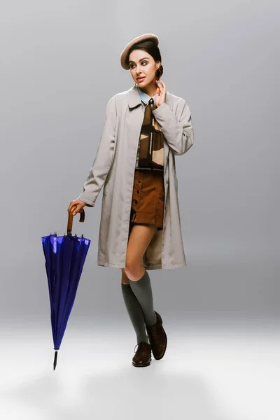 Full length of elegant young woman in beret and trench coat posing with blue umbrella on grey — Stock Photo