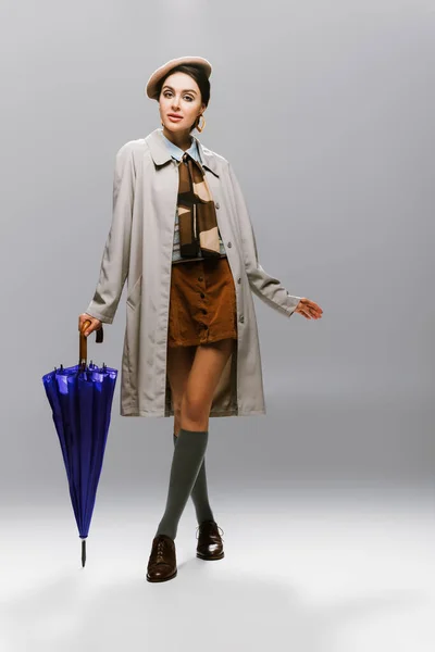 Full length of brunette young woman in beret and trench coat posing with blue umbrella on grey — Stock Photo