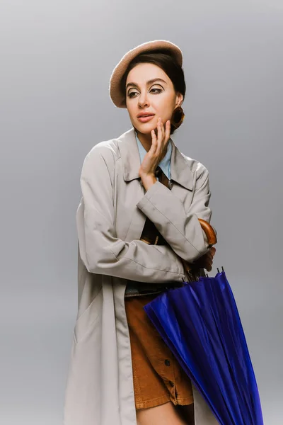 Brunette jeune femme en béret et élégant trench coat posant avec parapluie bleu isolé sur gris — Photo de stock
