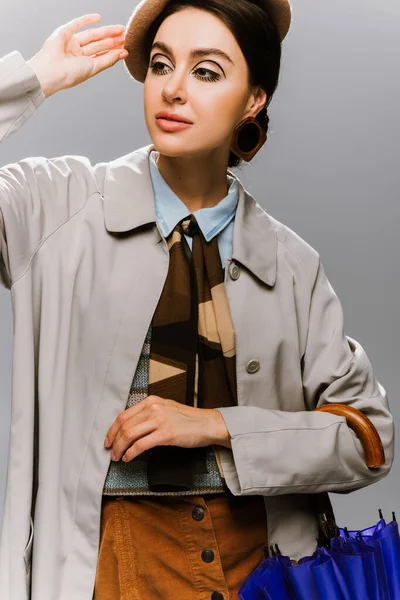 Retrato de una joven morena ajustando la boina y sosteniendo un paraguas azul aislado en gris - foto de stock