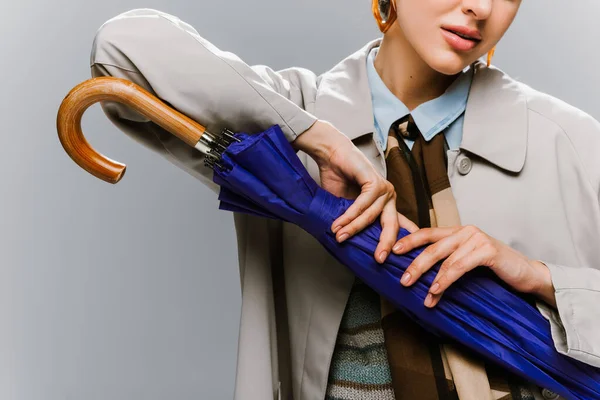 Cropped view of young woman in trench coat posing with blue umbrella isolated on grey — Stock Photo