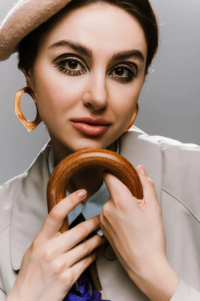 Retrato de mujer joven morena en boina y gabardina posando con paraguas aislado en gris - foto de stock