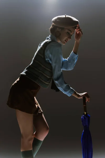 Silhouette de jeune femme en tenue automnale ajustant béret et debout avec parapluie sur gris foncé — Photo de stock