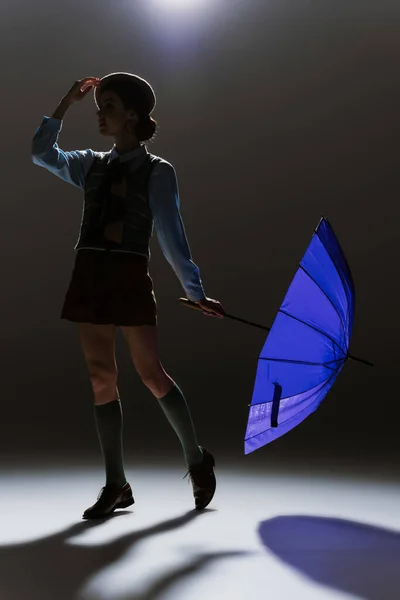 Силуэт элегантной молодой женщины в берете и юбке, позирующей с голубым зонтиком на сером — стоковое фото