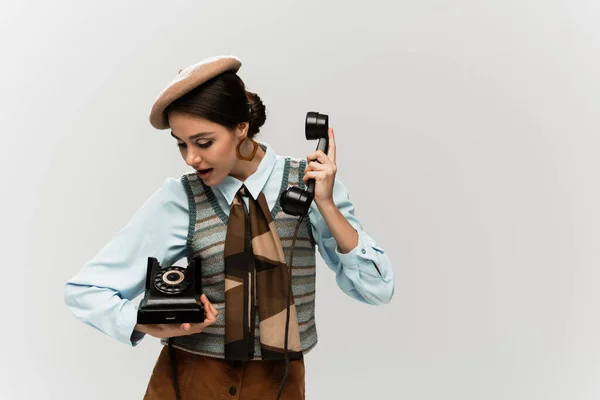 Mujer joven asombrada en boina sosteniendo teléfono vintage aislado en gris - foto de stock