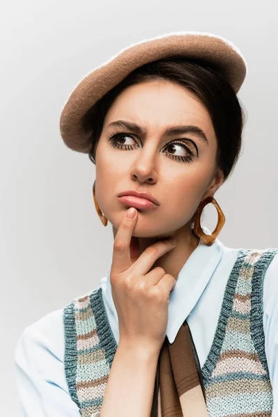 Mujer joven pensativa con maquillaje en boina y chaleco de punto aislado en gris - foto de stock