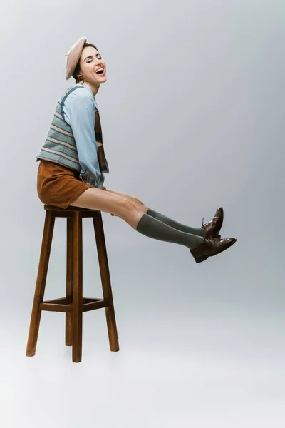 Full length of amazed young woman in beret and autumnal clothes sitting on wooden high chair on grey — Stock Photo