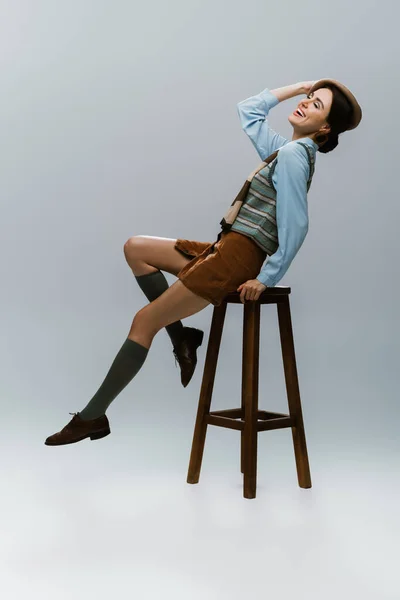 Full length of positive young woman in beret and autumnal clothes sitting on wooden high chair on grey — Stock Photo