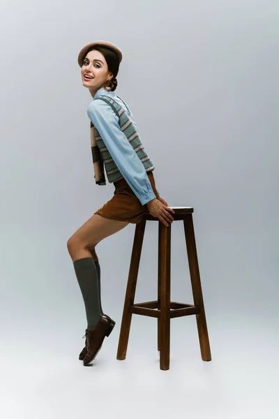 Full length of pleased young woman in beret and autumnal clothes sitting on wooden high chair on grey — Stock Photo