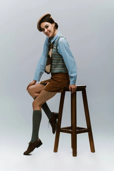 Full length of happy young woman in beret and autumnal clothes sitting on wooden high chair on grey — Stock Photo