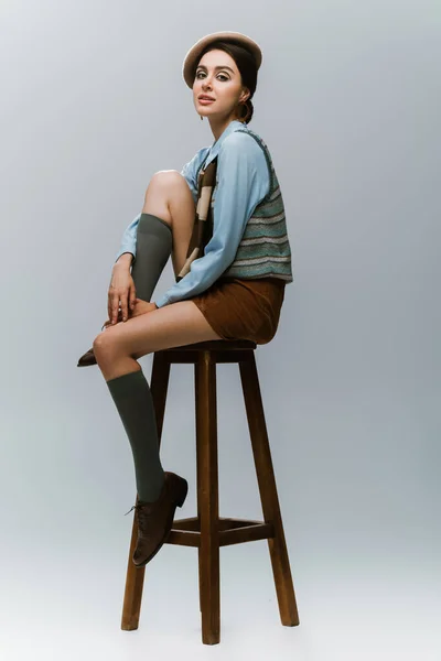 Longitud completa de la mujer joven en boina y ropa otoñal sentado en la silla alta de madera en gris - foto de stock
