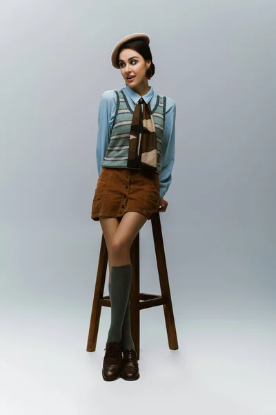 Full length of young woman in beret and autumnal clothes standing near wooden high chair on grey — Stock Photo