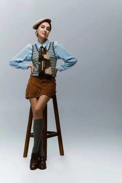 Volle Länge der jungen Frau in Baskenmütze und herbstlicher Kleidung stehend mit den Händen auf den Hüften in der Nähe von hölzernen Hochstuhl auf grau — Stockfoto