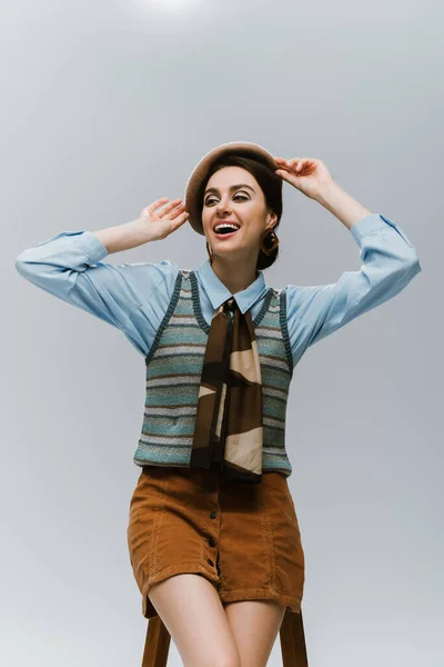 Amazed young woman in autumnal clothes adjusting beret near wooden high chair on grey — Stock Photo