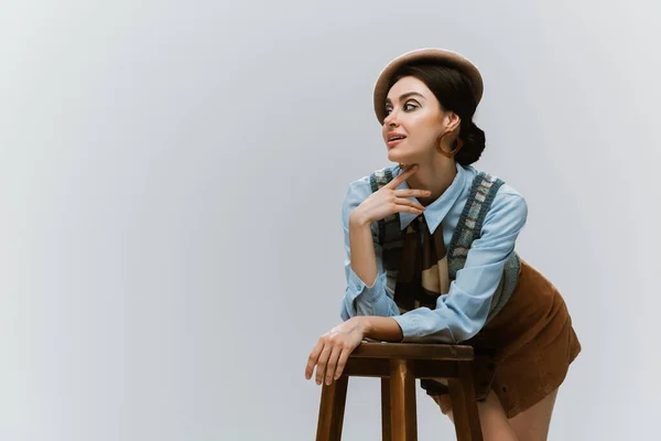 Jeune femme en béret et vêtements automnaux appuyé sur chaise haute en bois isolé sur gris — Photo de stock