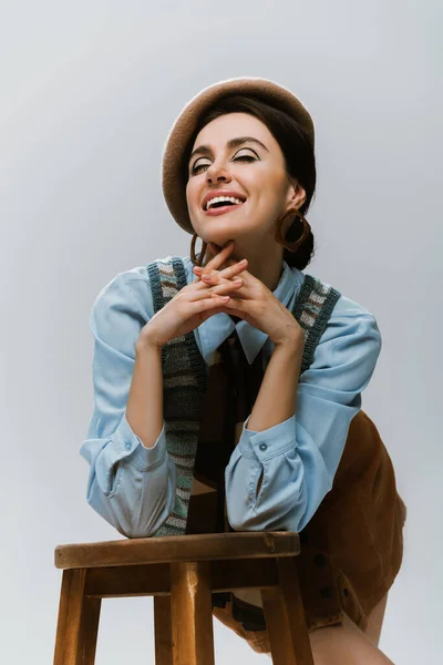 Cheerful young woman in beret and autumnal clothes leaning on wooden high chair on grey — Stock Photo