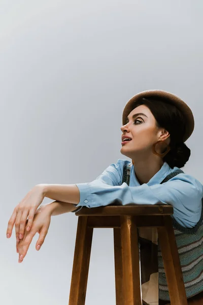 Young stylish woman in beret and autumnal clothes leaning on wooden high chair isolated on grey — Stock Photo