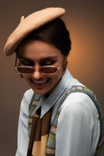 Happy young woman in beret and stylish sunglasses smiling on brown — Stock Photo