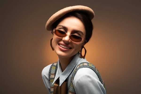 Веселая молодая женщина в берете и стильных солнцезащитных очках, улыбающаяся на коричневом — стоковое фото