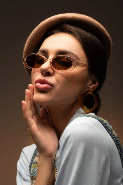 Élégant jeune femme en béret et lunettes de soleil boudant lèvres sur brun — Photo de stock