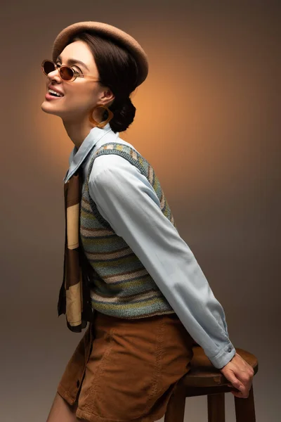 Smiling young woman in beret and sunglasses sitting on high chair on brown — Stock Photo