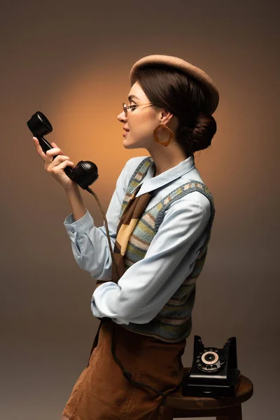 Élégant jeune femme en béret et lunettes de soleil en regardant le téléphone vintage sur brun — Photo de stock