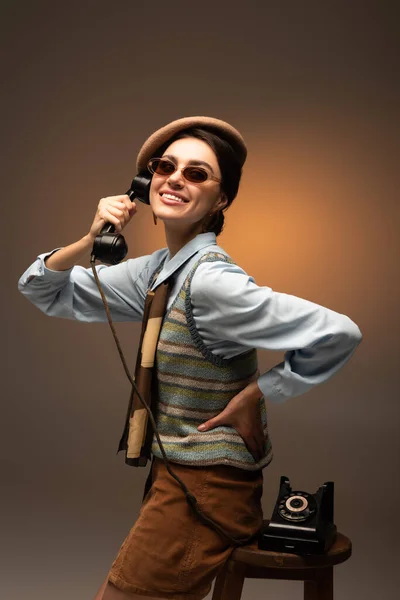 Fröhliche junge Frau in Baskenmütze und Sonnenbrille mit Vintage-Telefon auf braun — Stockfoto