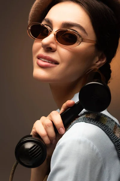 Stylish young woman in beret and sunglasses holding vintage telephone on brown — Stock Photo