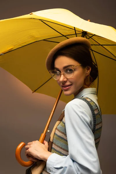 Joyful young woman in beret and eyeglasses holding yellow umbrella on grey — Stock Photo