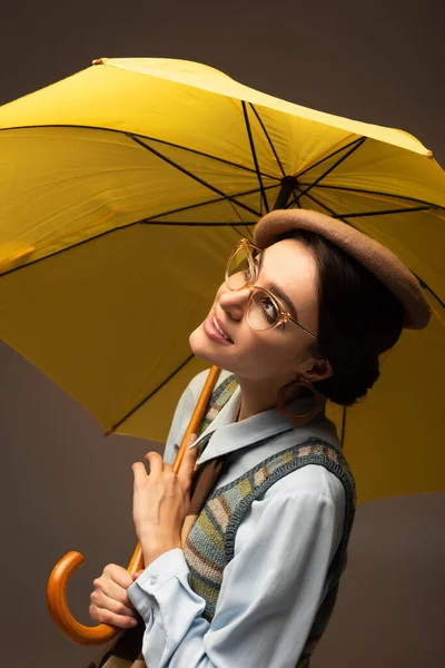 Positive young woman in beret and eyeglasses holding yellow umbrella on grey — Stock Photo
