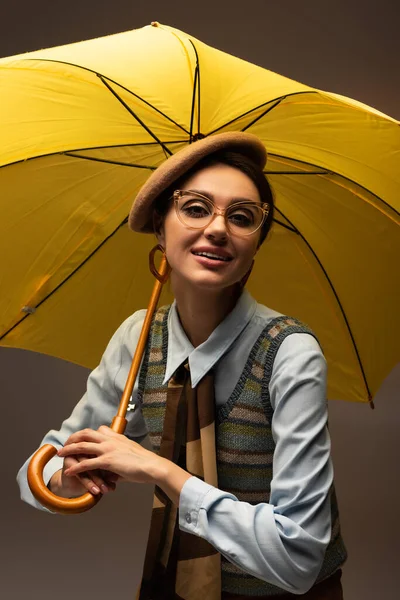 Joyeuse jeune femme en béret et lunettes tenant parapluie jaune sur gris — Photo de stock