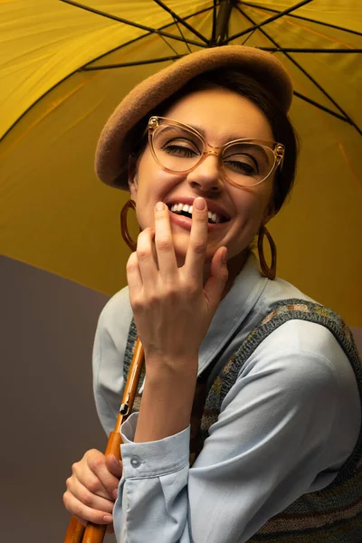 Feliz jovem mulher em boina e óculos segurando guarda-chuva amarelo no cinza — Fotografia de Stock